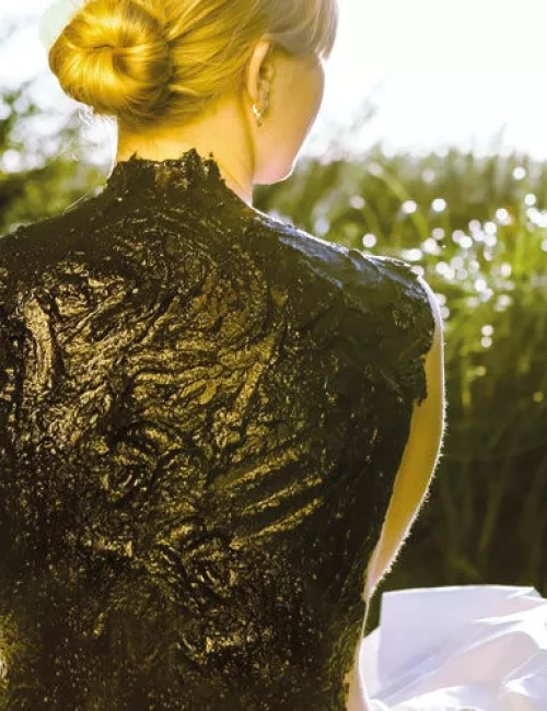 Eine Frau mit blonden Haaren in einem Dutt sitzt im Freien mit dem Rücken zur Kamera, ihr Rücken ist vollständig mit einer schwarzen Moorpackung bedeckt.