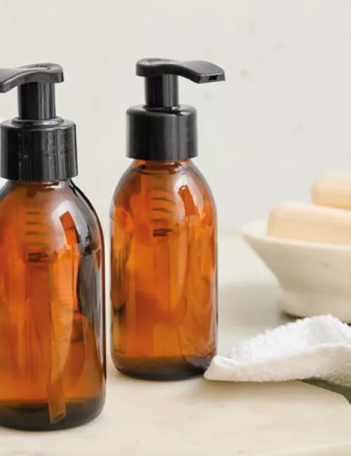 Zwei braune Spenderflaschen mit schwarzem Pumpverschluss stehen auf einem weißen Untergrund, daneben Handtücher, Eukalyptusblätter und eine Seifenschale.