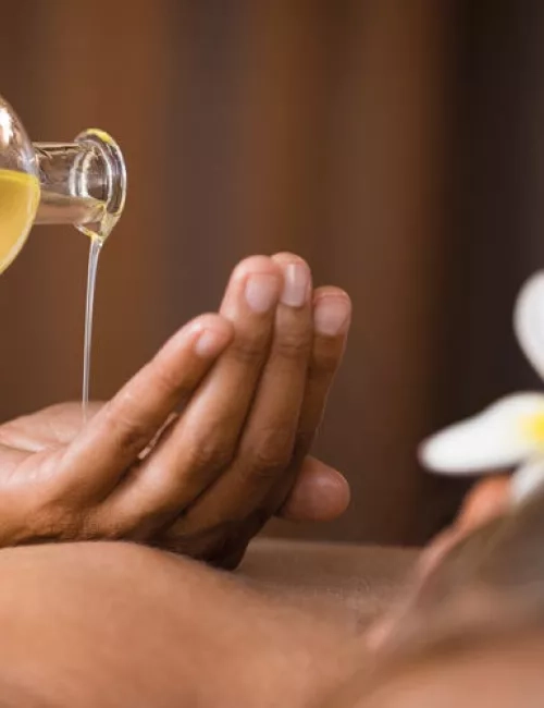 Warmes Massageöl wird in die Hände einer Person gegossen, im Vordergrund liegt eine entspannte Person mit einer Blume im Haar.
