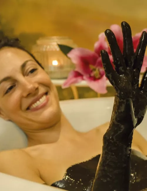 Frau entspannt in einer Badewanne und hält ihre Hand, die mit dunklem Schlamm bedeckt ist, hoch.