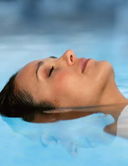 Eine Frau liegt entspannt auf dem Rücken im Wasser eines Pools und hat die Augen geschlossen. Ihr Gesicht ist teilweise unter Wasser, die Wasseroberfläche ist ruhig und spiegelt ihr Profil. Die Atmosphäre wirkt friedlich und erholsam.