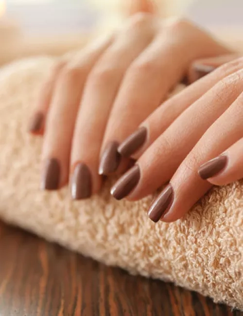 Nahaufnahme gepflegter Frauenhände mit braun lackierten Fingernägeln, die entspannt auf einem beigen Frotteehandtuch ruhen; im Hintergrund sorgen weiche Lichtreflexe für eine warme, entspannende Atmosphäre.