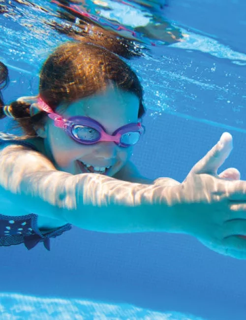Lachendes Mädchen mit rosa Schwimmbrille taucht unter Wasser in einem Pool und streckt die Arme nach vorne.