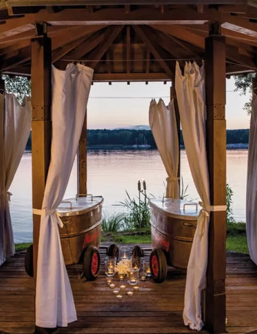 Romantischer Pavillon mit weißen Vorhängen und zwei nostalgischen Badewannen auf Holzplattform, beleuchtet von Kerzenlicht mit Blick auf einen ruhigen See bei Sonnenuntergang.