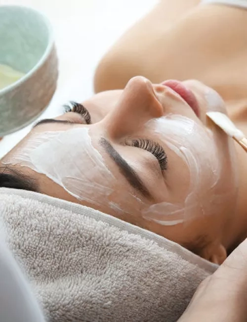 Eine Frau liegt entspannt mit geschlossenen Augen auf einer Liege, während ihr eine weiße Gesichtsmaske mit einem Pinsel aufgetragen wird. Sie ist in ein Handtuch eingehüllt, und eine Schale mit der Maske wird danebengehalten. Die Szene vermittelt eine ruhige Wellness-Atmosphäre.