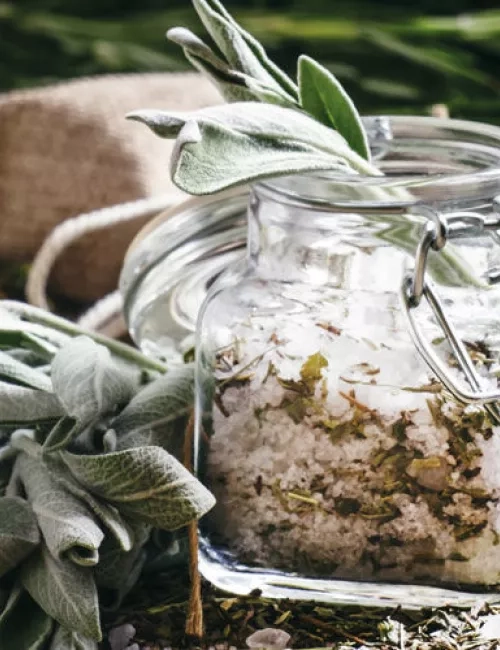 Ein Glas mit aromatisiertem Badesalz, gefüllt mit Kräutern und Meersalz, steht auf einem Holztisch, umgeben von frischen Salbeiblättern und getrockneten Pflanzenteilen.