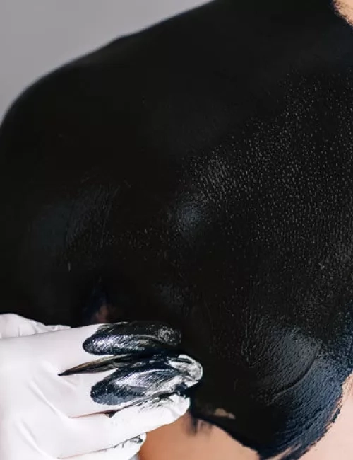 Eine Person trägt mit behandschuhten Händen eine dicke, schwarze Moorpackung auf den Rücken eines anderen Menschen auf.