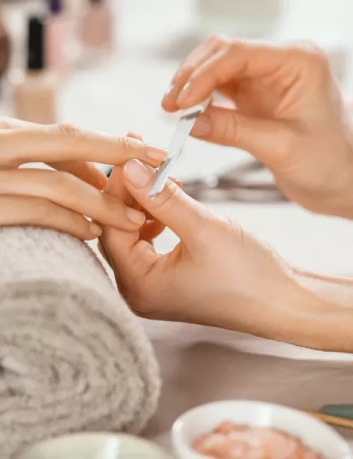 Nahaufnahme einer professionellen Maniküre: Eine Person feilt vorsichtig die Fingernägel einer anderen, die Hand liegt entspannt auf einem grauen Frotteehandtuch; im Vordergrund stehen kleine Schalen mit Pflegeprodukten.