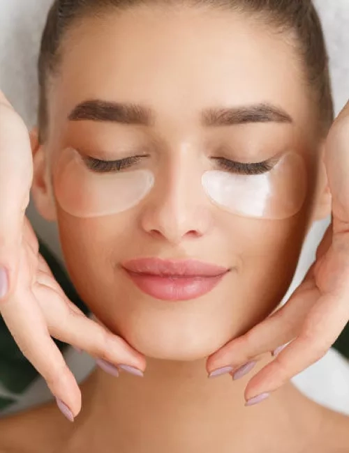 Eine junge Frau mit geschlossenen Augen liegt entspannt da, während transparente Augenpads unter ihren Augen haften. Sanfte Hände rahmen ihr Gesicht, das von gepflegten Augenbrauen und leichtem Make-up betont wird.