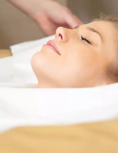 Eine entspannte Frau liegt mit geschlossenen Augen auf einer Behandlungsliege, eingehüllt in ein weißes Tuch, während eine Hand sanft ihren Kopf oder Nackenbereich berührt.