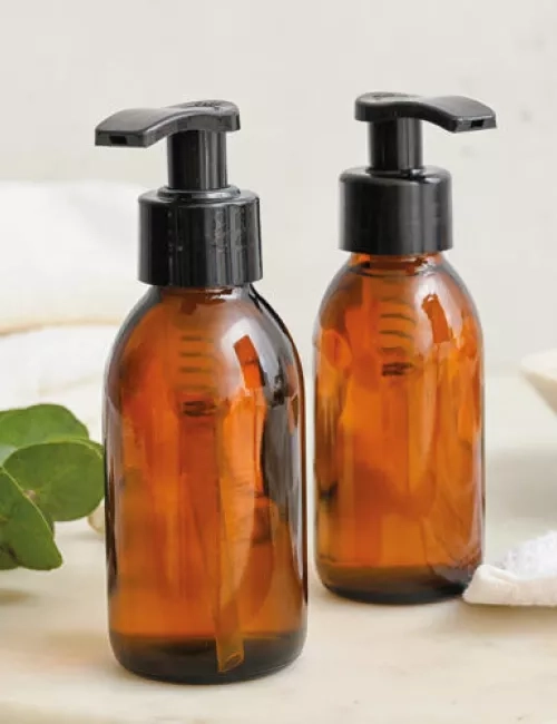 Zwei braune Spenderflaschen mit schwarzem Pumpverschluss stehen auf einem weißen Untergrund, daneben Handtücher, Eukalyptusblätter und eine Seifenschale.