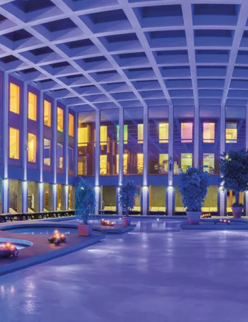 Abendlicher Innenbereich der SaarowTherme mit beleuchtetem Solebecken. Lila gefärbtes Wasser durch Unterwasserscheinwerfer, dekoriert mit Grünpflanzen und brennenden Kerzen am Beckenrand. Goldenes Licht fällt durch die Glasfront im oberen Bereich des Gebäudes.