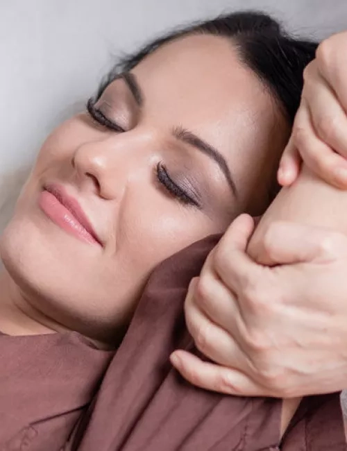 Zufriedene Frau mit geschlossenen Augen in brauner Wellnesskleidung genießt eine entspannende Armmassage in ruhiger Wohlfühlatmosphäre.