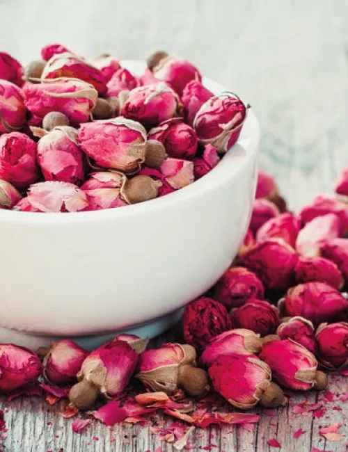 Eine weiße Schale gefüllt mit getrockneten Rosenknospen in kräftigem Pink, umgeben von weiteren Rosenknospen und Blütenblättern auf einem rustikalen Holztisch.