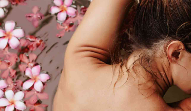 Wellnessbad in der SaarowTherme, Badewanne gefüllt mit Wasser, auf dem Wasser treibende Blumen in hellrosa und dunkelrosa. Eine junge Frau, mit dunkelbraunen Haaren und hellen Spitzen lehnt sich mit ihren Armen auf den Badewannenrand, sodass man den nicht bekleideten Rücken sieht. Der Kopf ist nach rechts gedreht und die Augen sind geschlossen.