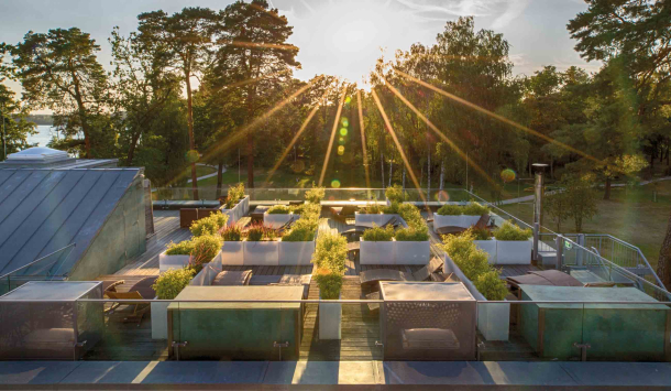 Die Dachterrasse der PanoramaSauna aus der Vogelperspektive fotografiert. Liegen mit braunen Auflagen unterschiedlich ausgerichtet und mit weißen Hüfthohen Blumenkübeln mit grünen Pflanzen in einzelne Komfortecken umrandet. Im Hintergrund sieht man grüne Bäume vom Kurpark,  den Scharmützelsee mit seinem blauen Wasser und die untergehende Sonne, die zwischen den Bäumen ihre Strahlen am blauen Himmel zeigt.