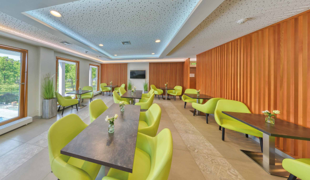 Ein großzügiger Raum mit senkrechten. mittelbraunen Holzverkleidungen als Säulen. Verteilt sind zahlreiche quadratische Tische mit dunkelgrauen Marmorplatten und grünen Sesselartigen Stühlen. Dekorative Akzente werden durch weiße Rosen in Vasen auf den Tischen gesetzt und grünen Stehpflanzen im Raum. Die Helligkeit erhält der Raum durch drei Bodentiefe Fenstern und den Deckenspots. An einer Wand hängt ein schwarzer Fernseher.