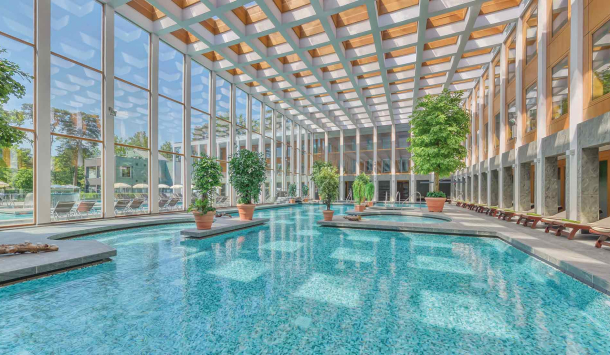 Thermalsolebad Innenbereich Tageslichtdurchflutet, türkisfarbenes Wasser. Dekorativ sind Grünpflanzen in verschiedenen Größen im Badbereich verteilt. Zahlreiche Liegemöglichkeiten mit grünen Decken und eine große Glasfront durch die man das Außenthermalsolebecken, Liegen und Sonnenschirme sieht. 