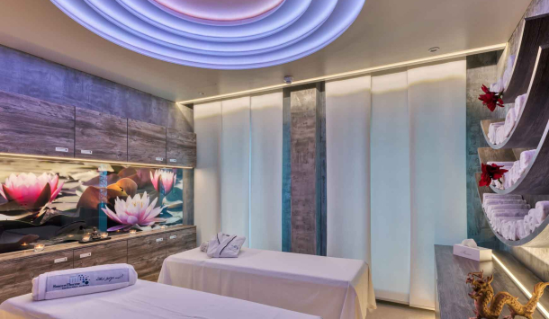 Ein Anwendungsraum mit zwei Massageliegen die mit weißen Laken bedeckt sind. Dekorativ befindet sich auf der einen Liege ein weißes Handtuch mit dem SaarowTherme Logo in Blau und auf der anderen ein weißer Bademantel mit Logo. Im Hintergrund sind graumelierte Hängeschränke mit einem Dekorativen Bild mit rosafarbenen Lotusblüten. Der Raum wird durch Deckenbeleuchtung und Unterschrankbeleuchtung warm weiß erleuchtet. Der Boden besteht aus einem Cremefarbenen Teppich. 