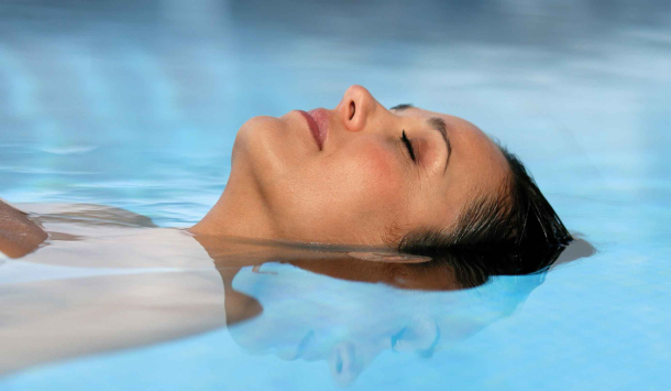 Eine Frau liegt entspannt auf dem Rücken im Wasser eines Pools und hat die Augen geschlossen. Ihr Gesicht ist teilweise unter Wasser, die Wasseroberfläche ist ruhig und spiegelt ihr Profil. Die Atmosphäre wirkt friedlich und erholsam.