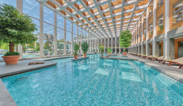 Thermalsolebad Innenbereich: Tageslicht, türkisfarbenes Wasser, Grünpflanzen und Liegen. Große Glasfront mit Blick ins Freie.