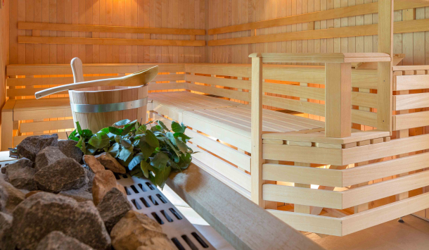 Hiki Sauna ausgestattet mit hellen Holz Sitzflächen und heller Holzwandverkleidung. Der Raum ist mit warm weißen Spots beleuchtet. Im Vordergrund des Bildes  befindet ein hölzerner Aufgusskübel mit einem großen Holzlöffel. Dieser befindet sich auf dem Saunaofen und als Dekoratives Element und Farbtupfer befinden sich einige braune Zweige mit grünen Blättern daneben.