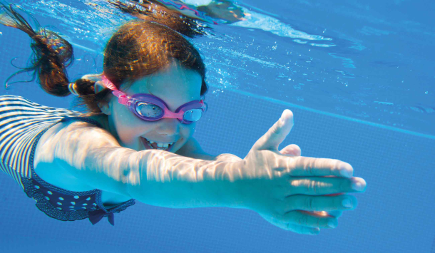 Mädchen mit Schwimmbrille taucht lachend unter Wasser und streckt die Arme nach vorn. Es trägt einen Badeanzug und schwimmt in einem hellblauen Schwimmbecken.
