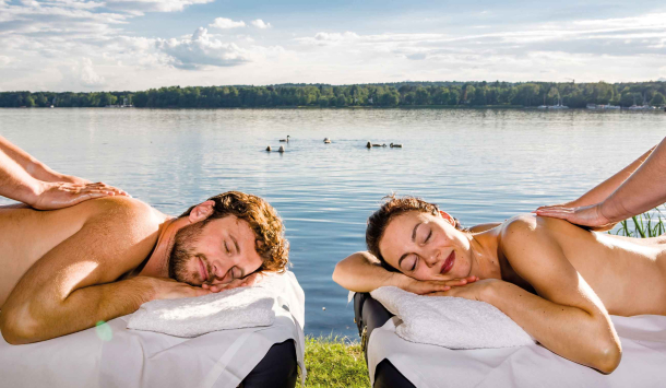 Ein Mann und eine Frau liegen sich auf zwei Massageliegen Kopf an Kopf gegenüber und werden direkt am See von zwei Therapeuten massiert. Die Augen der massierten Personen sind geschlossen und massiert werden die Rückenpartien. Im Hintergrund sieht man eine Schwanengruppe im Wasser, den glitzernden See und strahlenden Sonnenschein.