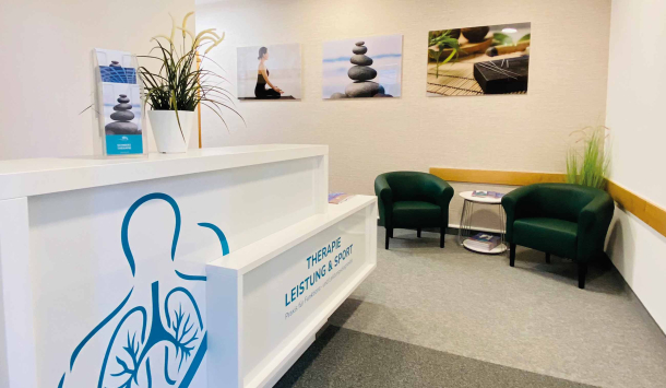 Zu sehen ist der Eingangsbereich der Arztpraxis an der SaarowTherme. Links vom Eingang befindet sich ein weißer Tresen mit Türkisfarbener Aufschrift "Therapie Leistung &Sport". Auf dem Tresen befindet eine kleine Grünpflanze und ein Prospektständer. Im Hintergrund befinden sich zwei dunkel grüne Sessel mit einem weißen, runden Couchtisch. Auf diesem befinden sich weitere Prospekte. Dekorativ ist der Eingangs Bereich mit einer Stehgrünpflanze und drei Bildern versehen. Hellgrauer Teppich, Cremefarbene Wand.