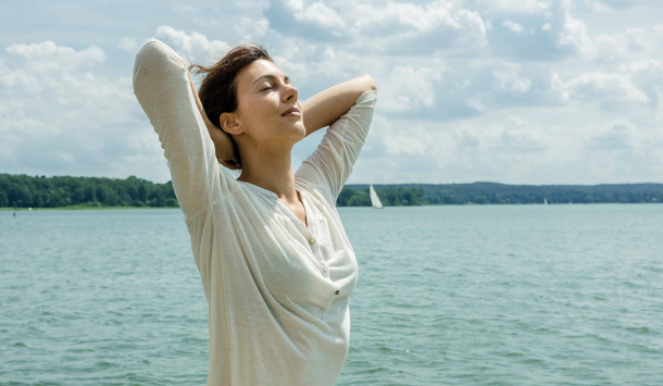 Eine Frau mit braunen Haaren und einem weißen langärmligen Shirt steht am See, verschränkt die Arme hinter dem Kopf und streckt ihr Gesicht in Richtung Sonne mit geschlossenen Augen. Im Hintergrund sind man Wald und davor den See mit Segelschiffen.