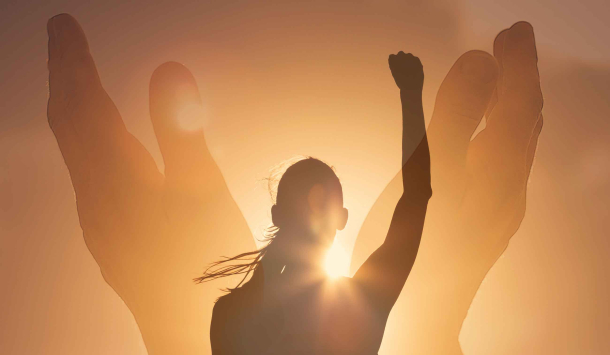 Ein Symbolbild, dass zeigt wie eine Frau die von der aufgehenden Sonne beschien wird und nur als Schatten zu erkennen ist. Sie hebt ihren rechten Arm mit einer geballten Faust in den Himmel. Als helleren Schatten sieht man zwei Hände die eine Art Schale um die Frau bilden. Durch die aufgehende Sonne wirkt das Bild Orange-Gelb. 