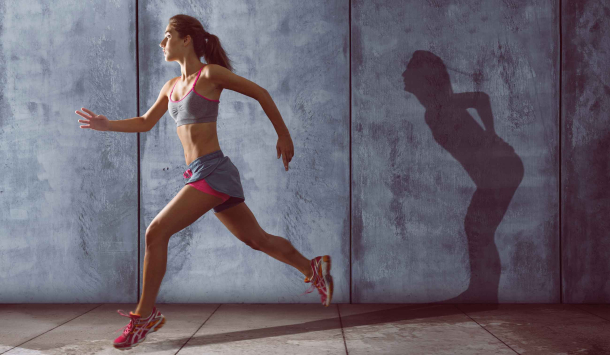 Eine trainierte Frau laufend vor einer grauen Wand in grauen, kurzen Sportsachen mit pinken Akzenten. Der Schatten wirft eine Frau die gebeugt ist und sich den unteren Rücken hält. 