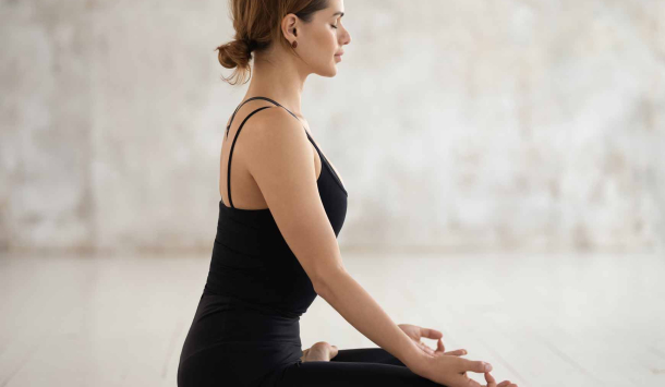 Eine Frau mit dunkelbraunen Haaren, einem Dutt, sitzt in einem schwarzen Yoga Outfit parallel zur Kamera in einem Schneidersitz mit geschlossenen Augen. Ihr Rücken ist sehr gerade und ihre Hände haben die Ohm Position eingenommen. Der Hintergrund ist hell  mit etwas dunkleren verwaschenen Spots. Sitzen tut die Frau auf einer schwarzen Yoga-Matte.