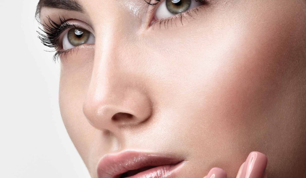Nahaufnahme eines weiblichen Gesichts mit gepflegter Haut, betonten Augenbrauen, langen Wimpern und zartem Make-up. Eine Hand mit lackierten Fingernägeln berührt das Kinn.