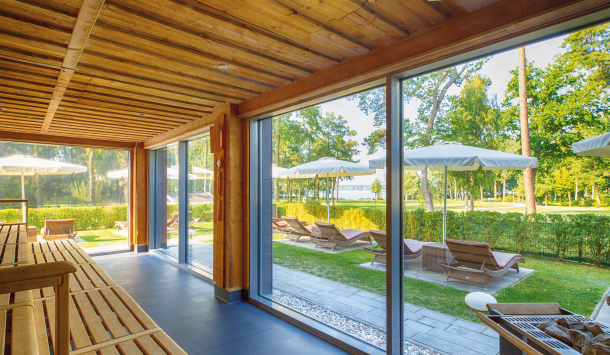 Abgebildet ist die KeloSauna der PanoramaSauna mit Blick auf den grünen Kurpark in Bad Saarow, mit Blick auf den blauen Scharmützelsee. Die Saunadecke und die Saunabank, sind mit groben Holzstrukturen versehen. Der Fußboden besteht aus dunkel grauen Fließen. Eine riesige Glasfront lässt es zu, dass der Raum Sonnendurchflutet wird und man sieht die braunen Außenliegen mit weißen aufgespannten Sonnenschirmen. Ein dunkel grauer Saunaaufgussofen ist am Rande des Bildes zu erkennen.