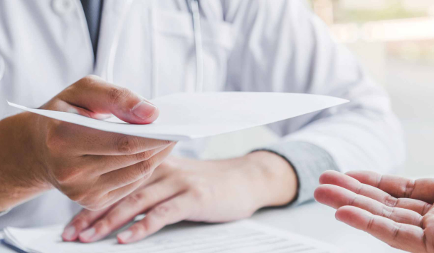 Ein Arzt in weißem Kittel überreicht einem Patienten ein Dokument, vermutlich ein Rezept oder Befund. Nur die Hände der beiden Personen sind sichtbar.