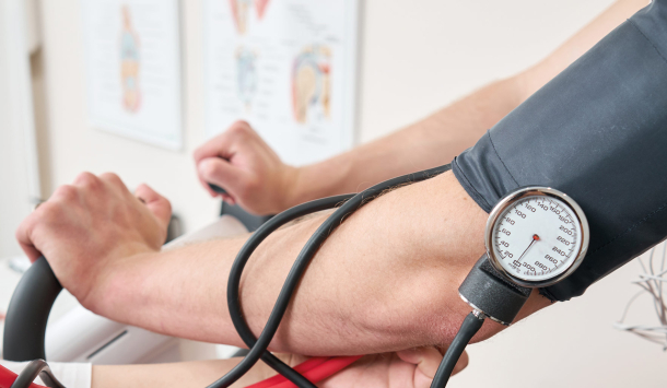 Ein Man sitzt auf einem Sportrad in einer Arztpraxis und macht einen Leistungstest. Ein Arzt steht an seiner Seite und hält ein Stethoskop an seinem Arm und messt gleichzeitig sein Blutdruck. Im Hintergrund erkennt man die Einrichtung einer Arztpraxis.