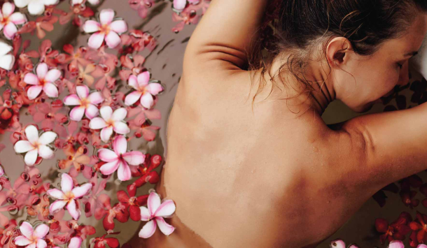 Frau liegt in Bauchlage leicht angelehnt am Rand einer Badewanne. Die Badewanne ist mit Blütenwasser gefüllt. Die Frau genießt ein erholendes Wellnessbad.