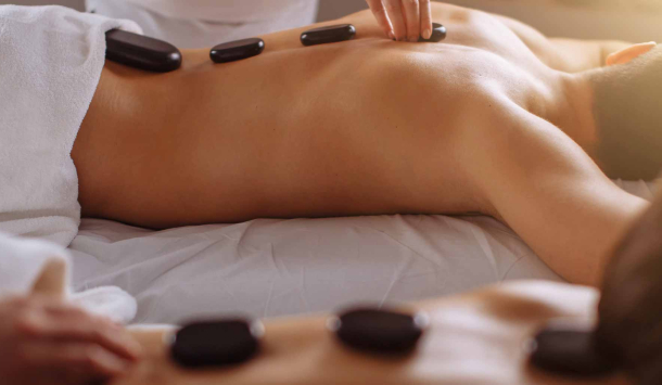 Ein Pärchen erhält eine HotStone Massage. Im Vorderund liegt eine Frau in Bauchlage auf einer Massageliege und dahinter sieht man einen Mann in gleicher Lage. Beide bekommen eine Massage mit warmen schwarzen Steinen.