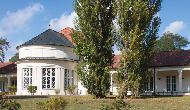 Das Bild zeigt ein elegantes, weißes Gebäude mit rundem Vorbau und roten Ziegeldächern, das sich inmitten einer gepflegten Gartenanlage befindet. Hohe Bäume und blühende Sträucher umrahmen das Gebäude, der Himmel ist blau mit wenigen Wolken.