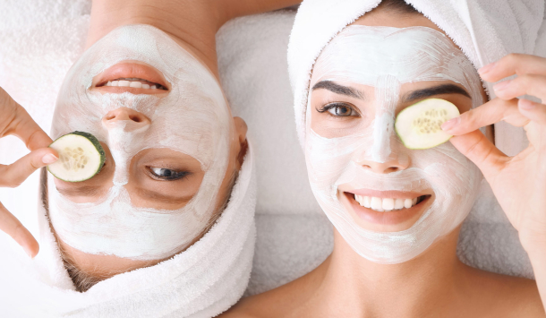 Zwei junge Frauen liegen auf dem Rücken auf einem Bett. Haare sind eingehüllt in einem Handtuch und auf dem Gesicht tragen Sie eine Gesichtsmaske und halten sich jeweils eine frische Gurke vor dem Auge.