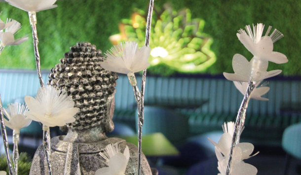 Aufnahme in einem asiatischen Restaurant. Im Vordergrund des Bildes steht eine kleine Buddha Figur mit weißen Lotusblüten. Im Hintergrund sieht man eine lange Sitzbank mit türkisblauem Stoff bezogen und an der Rückwand, die mit künstlichen Moos dekoriert ist, hängt eine aus Metall beschnittene Lotusform mit LED Hinterbeleuchtung.