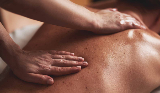 Frau liegt in Bauchlage auf eine Massageliege. Ihre Haare sind leicht zusammengesteckt und der Rücken mit Massageöl eingecremt. Eine Masseurin gibt ihr gerade eine Rückenmassage. 