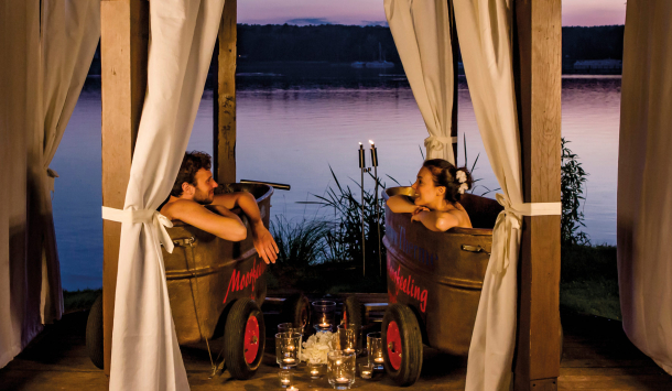 Junges Pärchen genießt ein Moorbad in einem romantischen Pavillon am See. Das Pärchen sitzt jeweilig in einer Wanne mit Moor befüllt. Sie schauen sich gegenseitig an und lächeln dabei. Auf dem Boden stehen ganz viele leuchtenden Teelichter und im Hintergrund erkennt man einen ruhigen See. Ein blau lila Farbspiel spiegelt sich auf der Wasseroberfläche vom See.