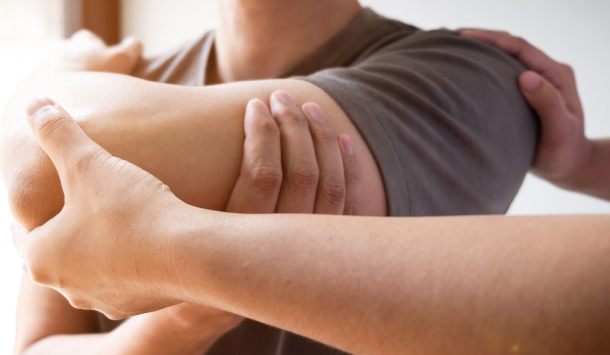 Ein Mann bekommt eine physiotherapeutische Behandlung. Mit einem Arm hält er den anderen Arm unter dem Ellbogen und rechts von ihm drückt ein Therapeut gegen seinen Ellbogen um den Schmerz zu lindern.