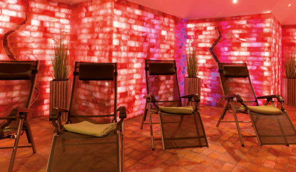 Mehrere Relaxliegen stehen in einem stimmungsvoll beleuchteten Ruheraum mit roten Salzwänden und warmem Licht. Der Boden ist mit Salzsteinen ausgelegt, an den Wänden stehen dekorative Pflanzen.