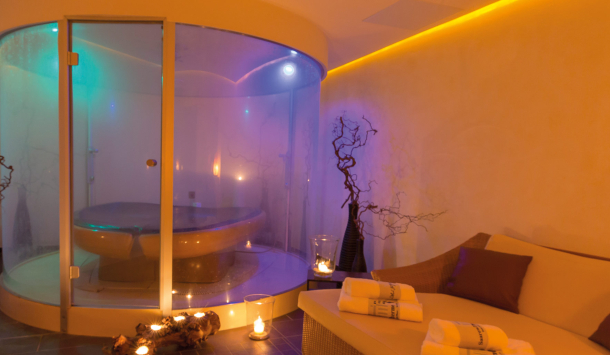 Das Salve-in-terra der SaarowTherme. Ein großer Raum mit in der Mitte eine Membranliege von einer Glaswand umgeben und in einem bläulichem Licht getaucht. Eine Duoliege mit Liegefläche zum Verweilen und Kerzenschein im Raum vermitteln eine romantische Atmosphäre.