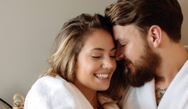 Junges Pärchen liegt auf einer Wellnesslounge mit kuscheligen Kissen in einem Beigeton. Das Paar umarmt sich und lächelt sich gegenseitig an. Sie tragen beide einen weißen Bademantel.