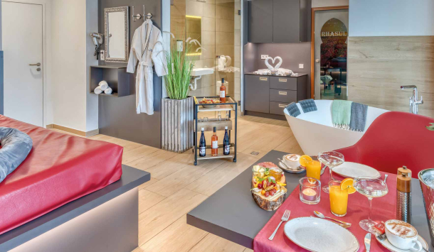 Ansicht des Private Spa der SaarowTherme. Im Vordergrund einen romantisch gedeckten Tisch mit Sektkühler, rote Rosen in einer hohen Vase und einem Sektfrühstück mit frischem Obst. Links im Bild erkannt man ein Wasserbett mit rotem Lederbezug. Im Raum sieht man einen großzügigen Duschbereich mit Toilette und Waschbeken. Rechts im Bild steht eine Duobadewanne und dahinter der Eingang zum eigenen Rhasul.