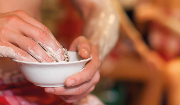 Ausschnitt von einer weißen Keramikschale mit Peelingmasse. Eine Männerhand nimmt gerade etwas Peelingcreme aus der Schale. Im Hintergrund sieht man zwei Frauen im Hamamtuch gekleidet, die sich auch mit Peelingcreme einreiben.